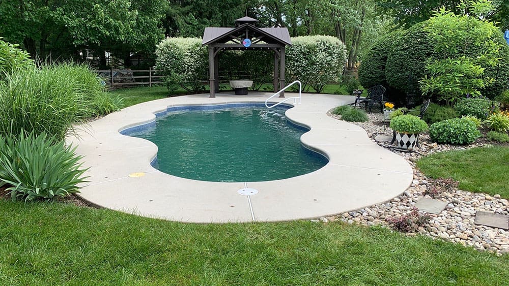 A small kidney-shaped pool with light blue water is surrounded by concrete decking and lush green grass. A wooden gazebo, perfect for a break from interior painting, sits at the far end, backed by dense shrubbery and trees. Various plants and bushes are arranged along the perimeter of the pool area.