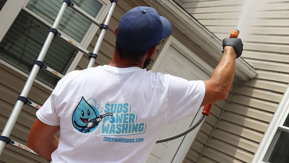 A person in a white T-shirt and blue cap is power washing the exterior of a house. The T-shirt has a logo with a smiling water drop holding a power washer, along with text reading "Suds Power Washing" and the website URL "justhiresuds.com." They also offer interior painting services to complete your home makeover.