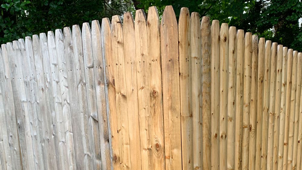 A wooden fence with vertical slats is shown, featuring one section painted gray and another in its natural wood color. This charming barrier sits against a lush green background filled with trees and foliage, reminiscent of an exterior that's as thoughtfully crafted as interior painting.