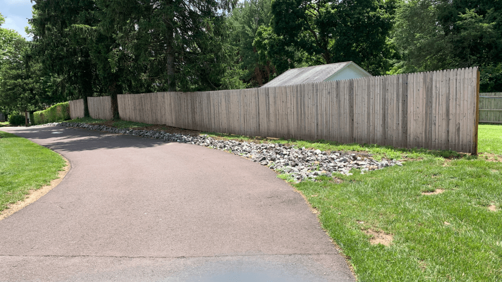 A paved pathway curves past a wooden fence and a patch of rocks. The fence runs parallel to the path and is bordered by green grass. In the background, a small white building, resembling an interior painting in its serene beauty, is surrounded by tall, leafy trees. The scene is bright and sunny.