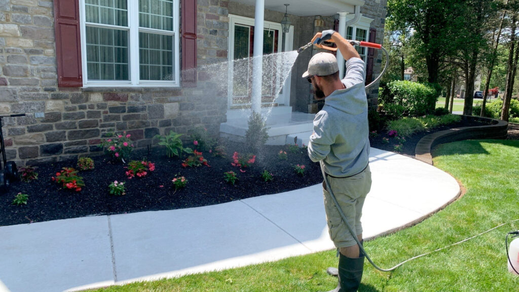 A person in a gray hoodie and cap is watering flowers in a garden bed near the entrance of a stone house. The garden bed, reminiscent of a Davis painting, boasts vibrant red and pink flowers. The curved pathway is neatly edged, and the lawn is lush and green.