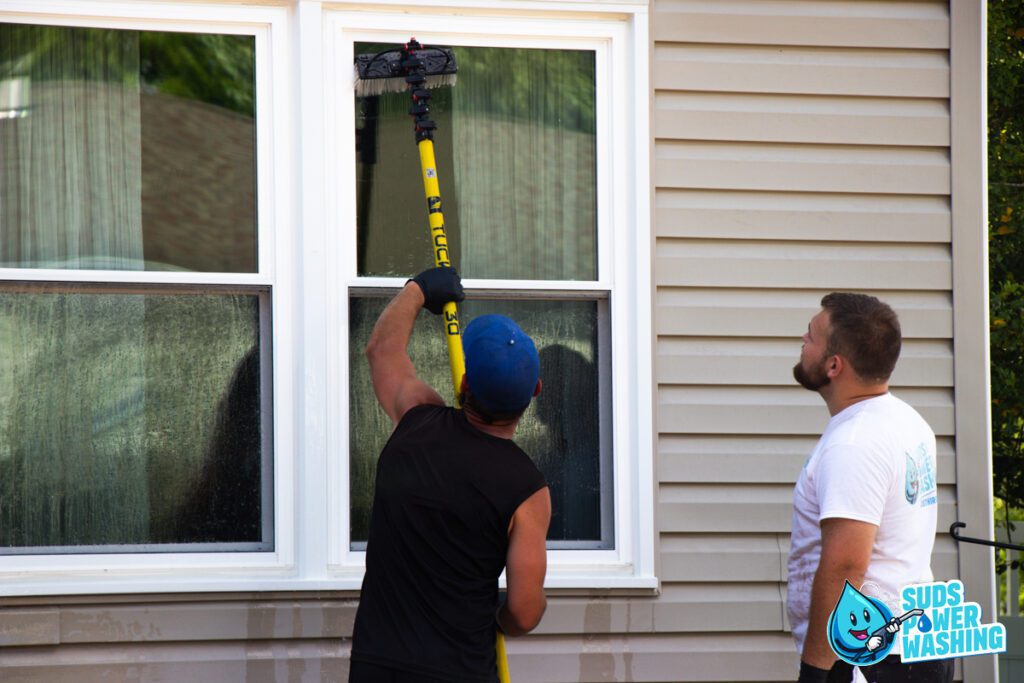 Two men are cleaning the exterior of a house. One man in a black tank top and blue hat uses a long brush attached to a pole to scrub a window, while the other man in a white shirt watches. The beige house with white-framed windows has the "Suds Power Washing" logo in the corner, perfect for your next social media post.