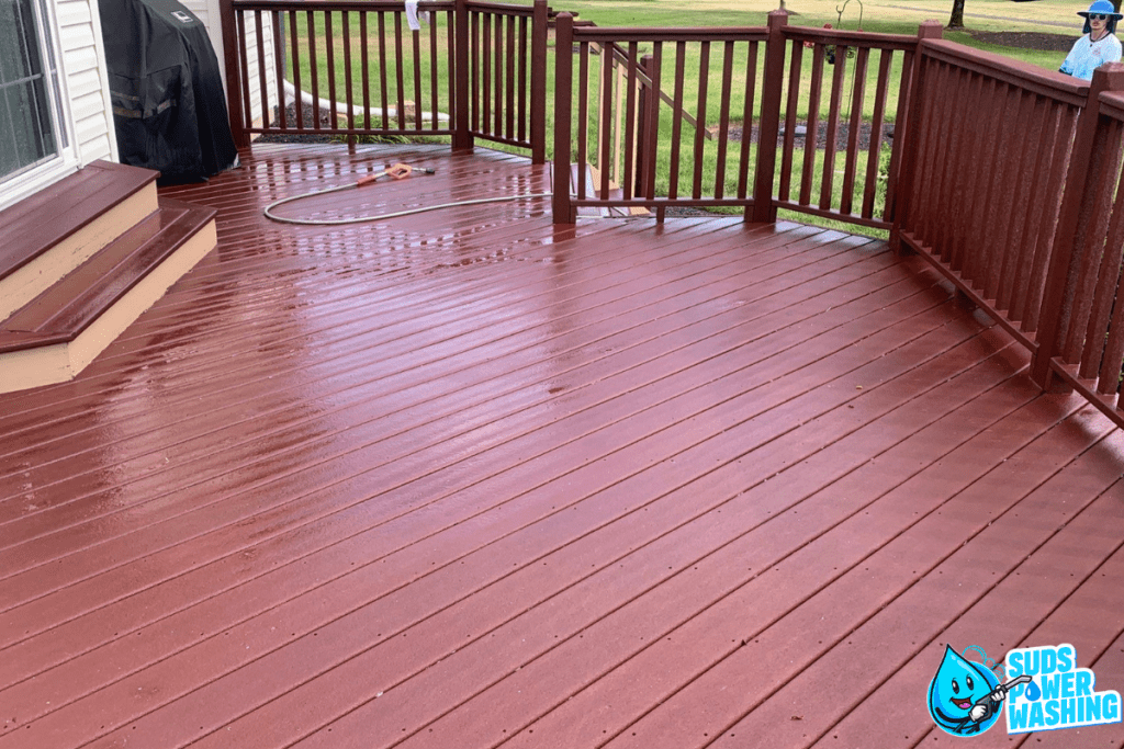 A freshly cleaned wooden deck with a hose coiled on it, surrounded by a brown railing. Steps lead down from a house on the left side, where clean gutters gleam in the sunlight. A grill is near the corner. The Suds Power Washing logo is at the bottom right corner. A person in a hat is in the background.