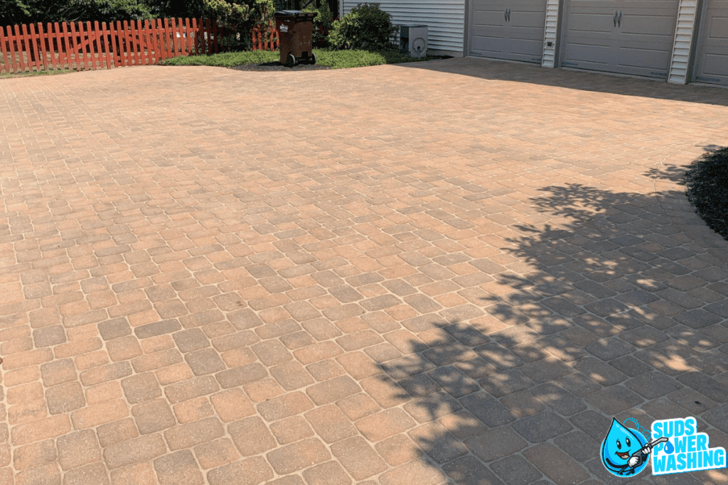 A clean, freshly power-washed brick driveway and concrete path in front of a garage with three closed doors. A red wooden fence and a trash bin are visible in the background, along with shadows from nearby trees. The Suds Power Washing logo is in the bottom-right corner.