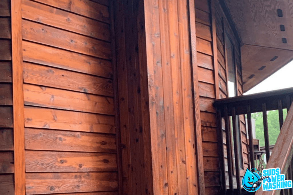 Close-up of a wooden house exterior with rich brown siding made from exotic woods and a corner post. A section of the roof with a rain gutter is visible, and a portion of a railing can be seen on the right. A logo for "Suds Power Washing" featuring a smiling water drop appears in the bottom right corner.