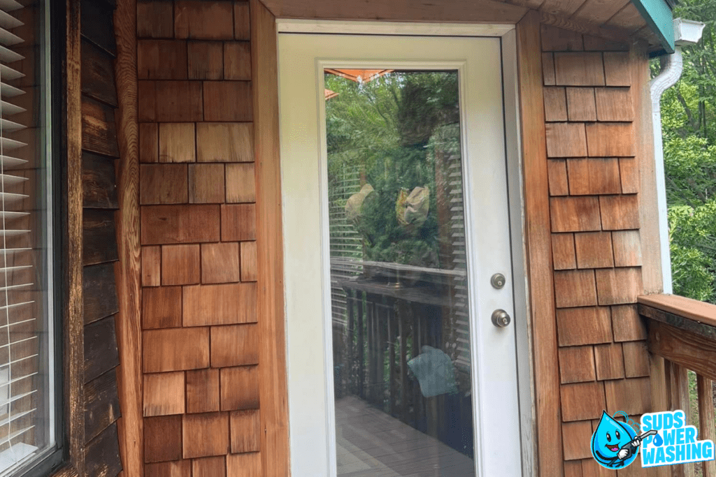 A wooden house exterior featuring a glass door with a white frame. The door's reflection shows green foliage and the house's porch railing crafted from exotic woods. To the right, a blue and white logo for "Suds Power Washing" with a smiling water drop character is visible.