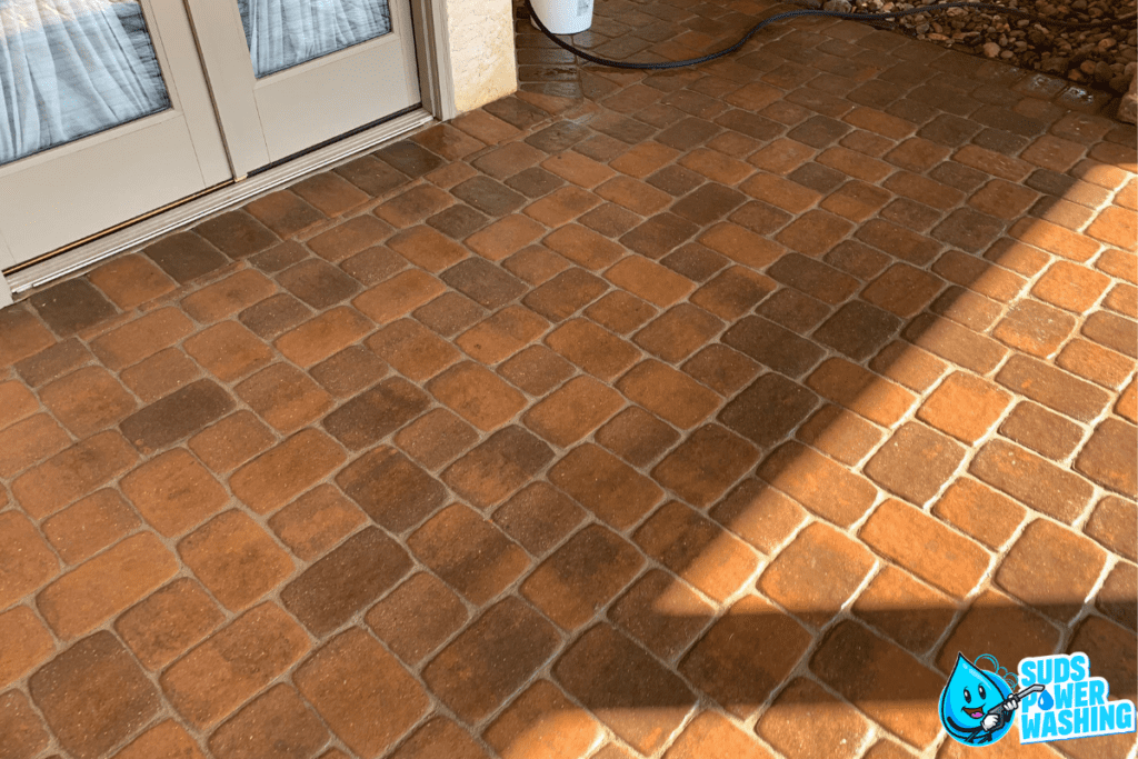 A sunlit outdoor brick patio features a clear power washing area marked by a distinct clean and dirty line. In the background, there’s a gray door and a bucket with a hose. The bottom right corner showcases the "Suds Power Washing" company logo with its blue bubble character.