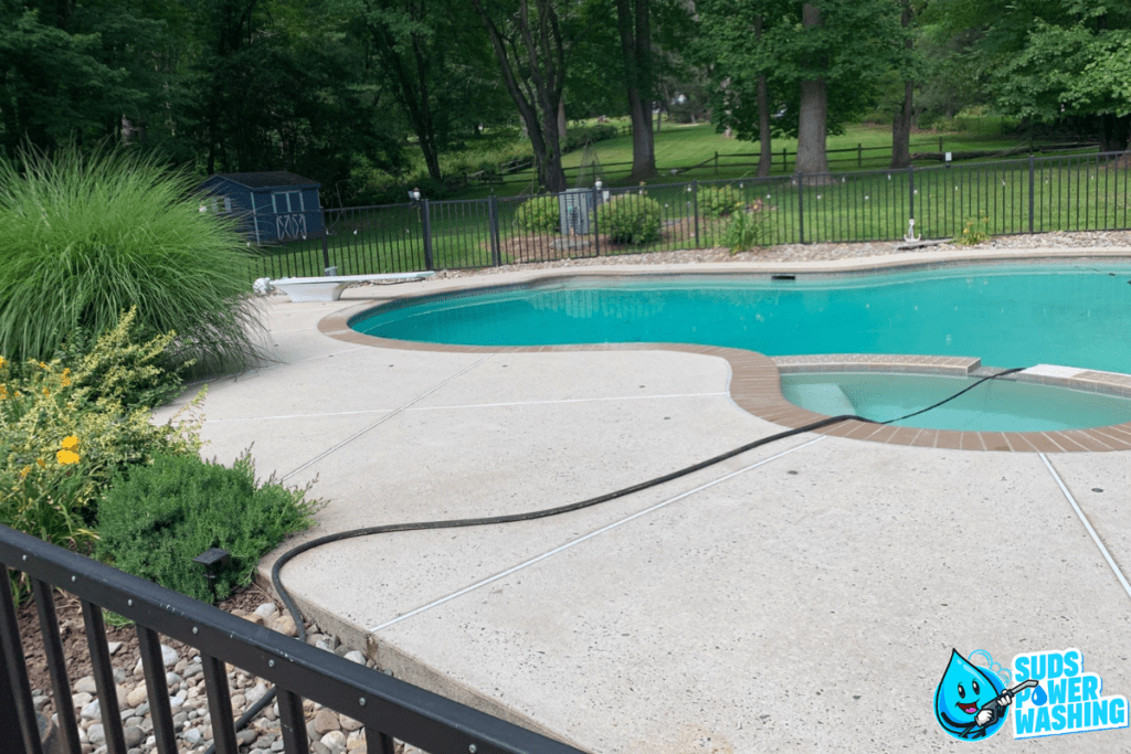 A clean swimming pool with clear water is surrounded by a light-colored concrete deck and walkways, bordered by greenery and a black fence. In the corner, a blue and white "Suds Power Washing" logo with a smiling water droplet is visible. Trees are in the background.