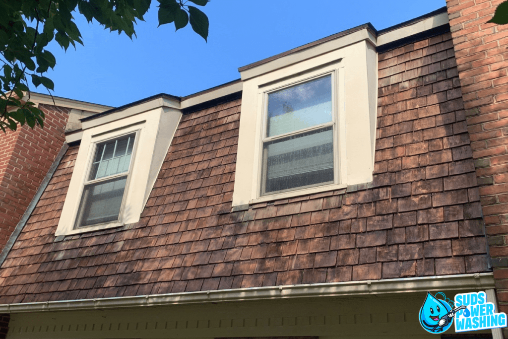 A close-up view of a house's upper level featuring two dormer windows with glass panes, framed by weathered brown shingles that mimic the texture of exotic woods. The sky is clear and blue, and a small company logo reading "Suds Power Washing" is visible in the bottom right corner.