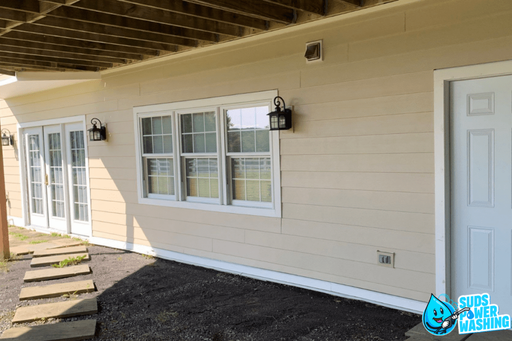 A beige exterior wall of a house with vinyl siding has two windows, each adorned with black outdoor lanterns. There is a white door on the right. A pathway of stone steps leads to the door. The logo "Suds Power Washing" featuring a water droplet character is in the lower right corner.