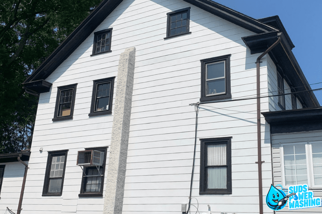 A two-story white house with black trim and vinyl siding is shown. The windows are framed in black, and there is an air conditioning unit in one window. A "Suds Power Washing" logo featuring a smiling water droplet mascot is in the bottom right corner of the image.