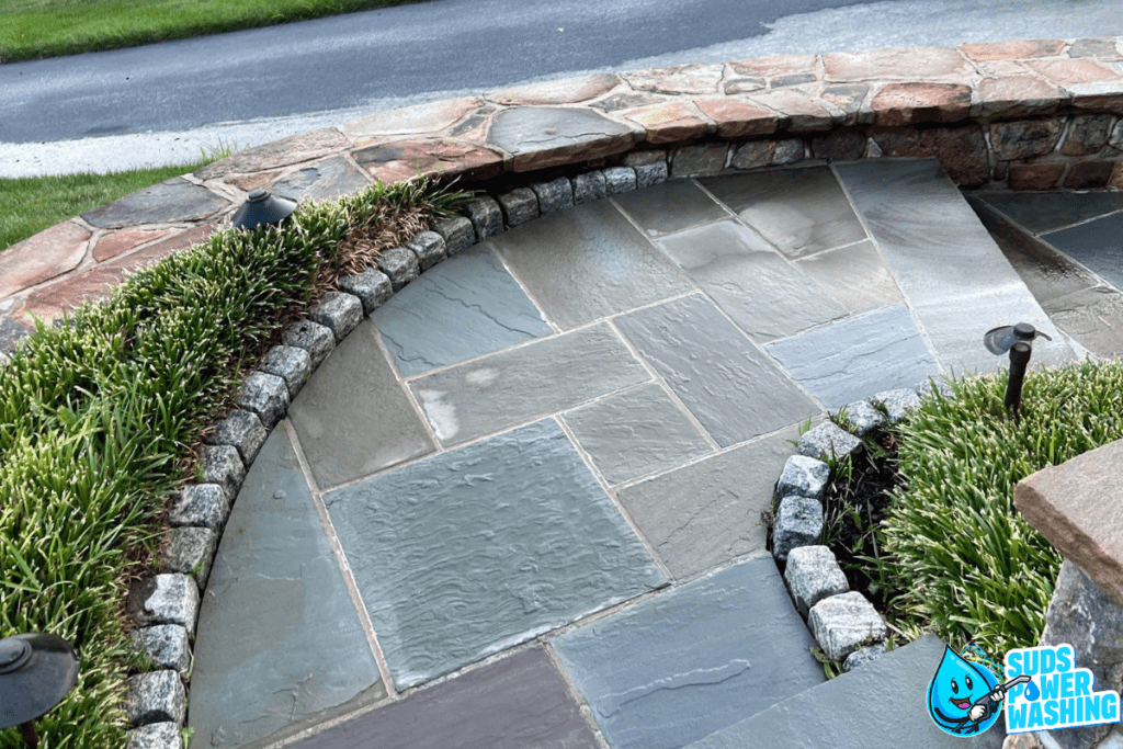 A curved stone pathway, bordered by cobblestones and greenery, leads to a stone-bordered area with lighting fixtures on either side. Composite decking subtly blends in, enhancing the setting. A road and grassy lawn are visible in the background. The Suds Power Washing logo is present in the bottom right corner.