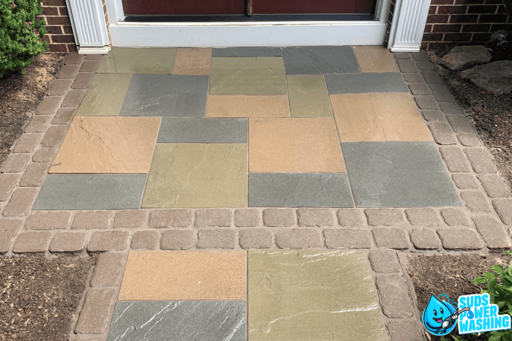 A freshly cleaned outdoor stone tile patio in front of a door. The patio, next to the composite decking, has a geometric pattern with grey, beige, and tan tiles framed by a stone border. The logo for Suds Power Washing featuring a smiling water drop is in the corner of the image.