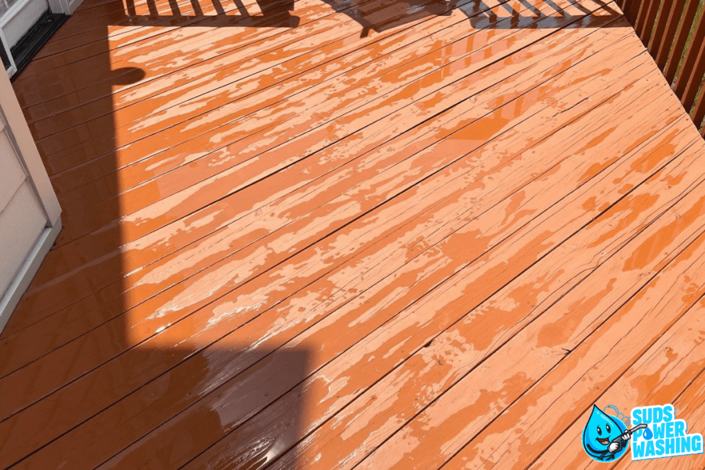 A freshly power-washed wooden deck, made from exotic woods, is shown drying in the sunlight. The deck boards are a light orangish-brown, and water droplets are scattered across the surface. In the bottom right corner, there's a logo for "Suds Power Washing" featuring a blue bubble character.