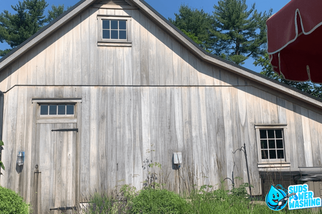 A weathered wooden building with two square windows and a door. The wood siding is surrounded by tall grass and greenery, while a red canopy is partially visible on the right side. The logo for "Suds Power Washing" is in the bottom right corner.