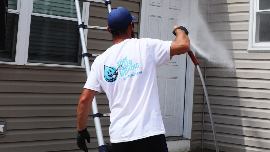 A person wearing a white "Sims Power Washing" t-shirt and a blue cap is using a pressure washer on the exterior of a house. A ladder propped against the wall, which displays a "Davis Painting" sticker, stands by a white door and window.