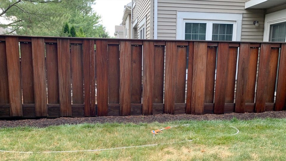 A tall, dark wooden fence stands in front of a residential building with beige siding and large windows. On the ground, there is a garden hose and a sprinkler lying on the grass, which is bordered by a mulch-covered flower bed along the fence. The pristine look owes its charm to Davis Painting's expert touch.