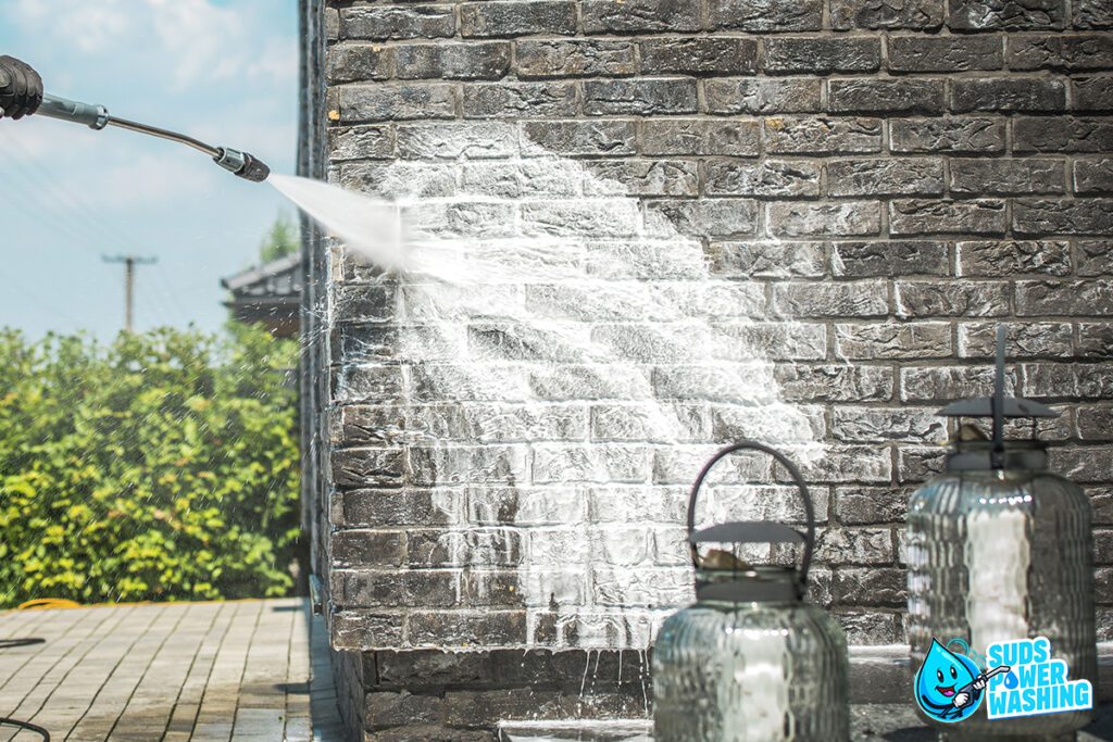 A high-pressure water stream from a power washer cleans a dark brick wall, creating a distinct, lighter area. Two large glass lanterns are positioned nearby, and greenery is visible in the background. Perfect prep work for any residential painting project. The Suds Power Washing logo is displayed in the bottom right corner.