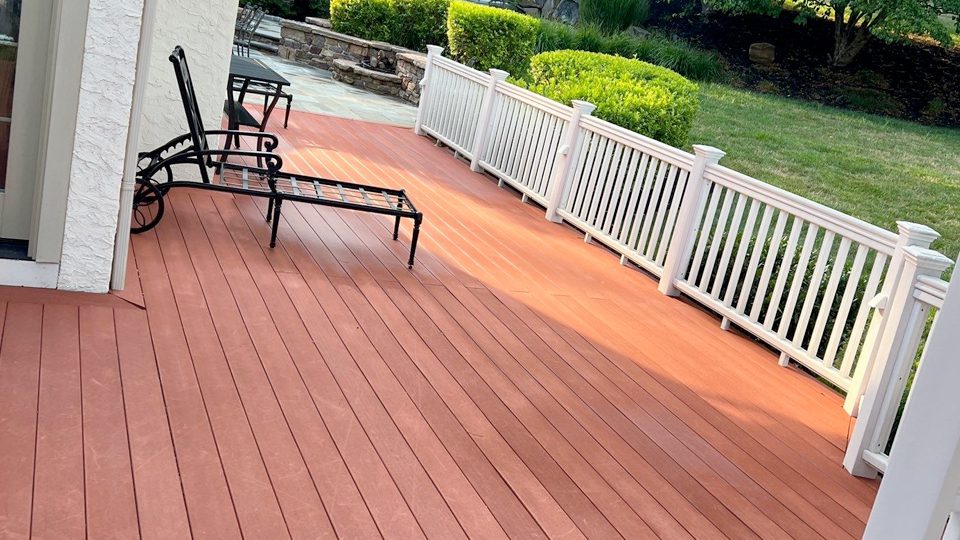 A wooden deck with reddish-brown planks is attached to a white building, courtesy of Davis Painting. The deck features white railings and two black metal recliners. In the background, there is a green lawn and a stone patio area with bushes and assorted plants.