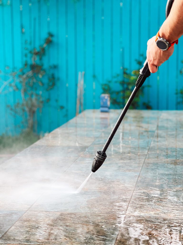 A person wearing an orange watch is power washing a tiled surface with a pressure washer, removing grime effortlessly. In the background, there is a blue fence and some plants are faintly visible. The scene gives off hints of recent residential painting activities.