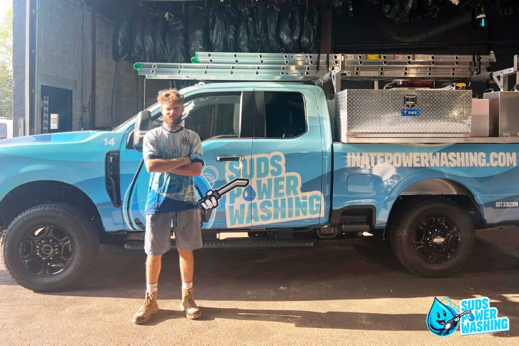 A man stands with his arms crossed in front of a blue truck that has "Suds Power Washing" and a website URL written on the side. The truck, essential to many careers in power washing, is equipped with tools and ladders. The man is wearing a casual outfit including a cap, T-shirt, shorts, and work boots.