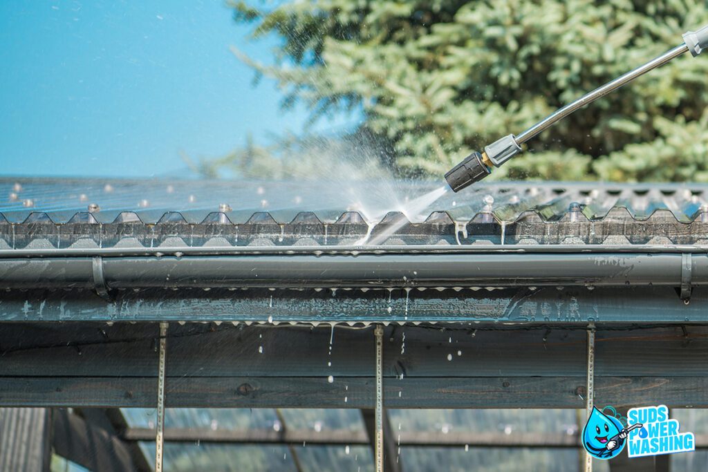 A pressure washing nozzle sprays water onto corrugated metal roofing under a clear blue sky, with clean windows glistening in the sunlight. The building appears to be an outdoor structure surrounded by green trees. The image includes the Suds Power Washing logo in the bottom right corner.