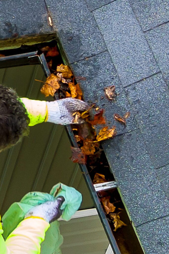 A person wearing a bright yellow jacket and white gloves uses a green cloth to clean out fallen autumn leaves from a roof gutter. The roof shingles, the edge of the gutter, and some nearby windows are visible. The brown leaves scattered around add to the seasonal feel.