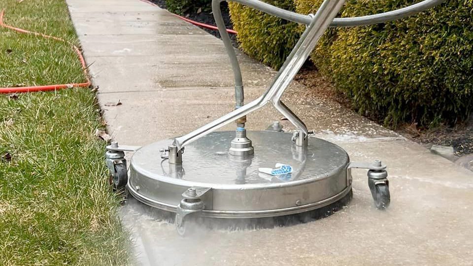 Close-up of a concrete sidewalk being cleaned by Davis Painting using a circular surface cleaner with multiple rotating nozzles attached to a handle. The cleaner is spraying water, creating a visible mist. Grass is on the left, and trimmed bushes are on the right.