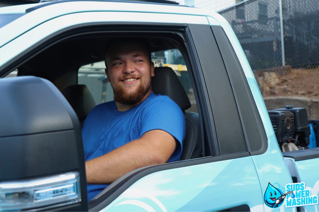 A smiling man with a short beard sits in the driver's seat of a truck bearing the Suds Power Washing logo. He is looking out the window, contemplating his rewarding career. The truck is light blue, and the background shows a chain-link fence and industrial buildings.