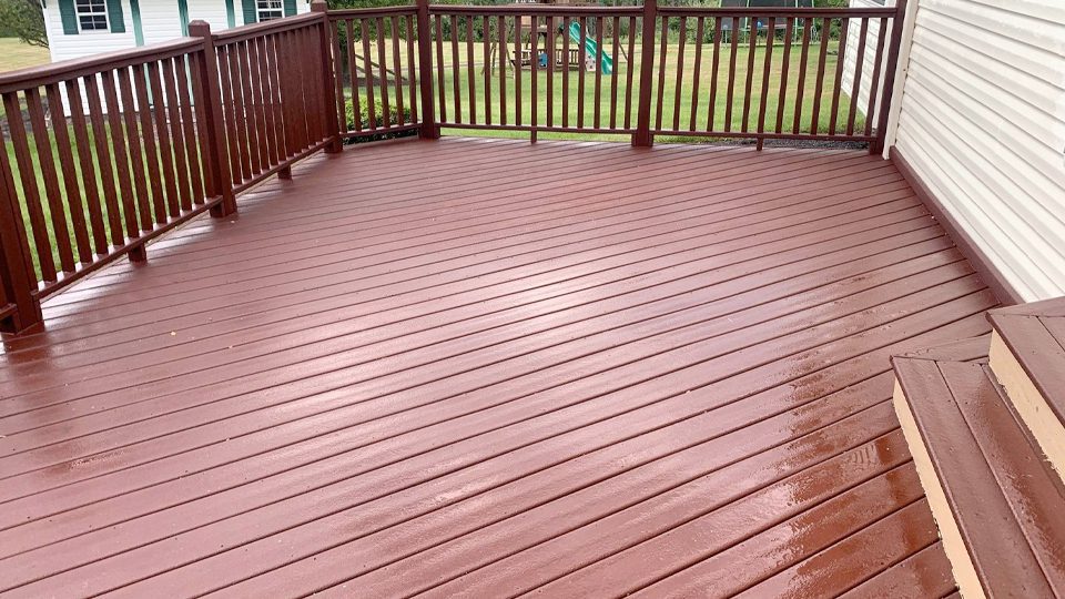 A freshly stained wooden deck with horizontal boards and red-brown railing, expertly done by Davis Painting, is adjacent to a white house and overlooks a grassy backyard with children's play equipment in the distance. The surface appears wet from rain or cleaning.