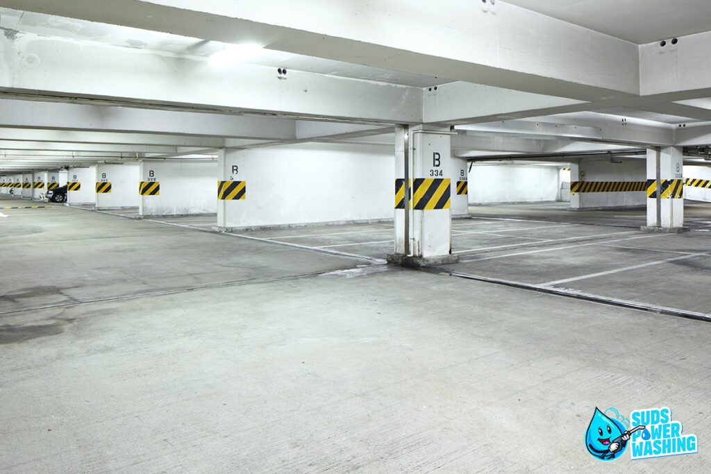 An empty underground parking garage. White support pillars are marked with yellow and black hazard stripes. The floor is clean, and the area is well-lit with fluorescent lighting. A "Suds Power Washing" logo featuring a cartoon soap bubble sits in the lower-right corner, hinting at its residential painting services.