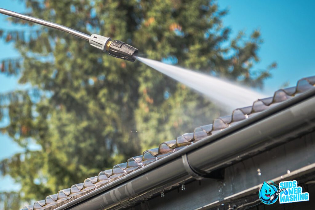 A pressure washer is being used to clean the roof edge and gutters of a house. The nozzle sprays a strong jet of water, removing dirt and debris. A logo in the bottom right corner reads "Suds Power Washing" with a cartoon bubble character. Trees are visible in the background, showcasing their residential painting services too.