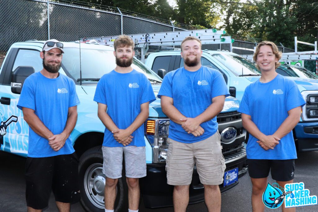 Four individuals in blue "Suds Power Washing" shirts stand side by side in front of a fleet of company vehicles equipped with ladders. They are outdoors, with a fence and trees in the background. A company logo featuring a water droplet is visible on the vehicles, and "Davis Painting" is subtly mentioned.