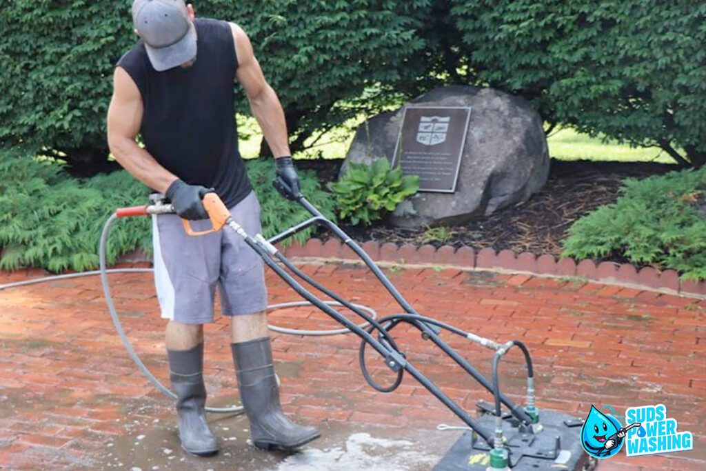 A person wearing a grey cap, black sleeveless shirt, grey shorts, gloves, and tall boots operates a pressure washing machine on a brick pathway. Surrounded by greenery and a sign on a rock in the background, the scene captures the essence of careers in outdoor maintenance. The "Suds Power Washing" logo is in the bottom right corner.