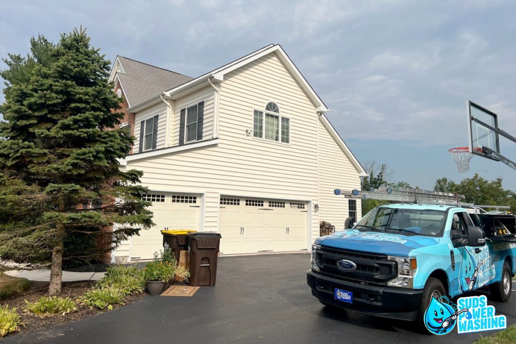 A white two-story suburban house with a large driveway and garage. Near the garage, there are two garbage bins. A blue work truck with cleaning equipment and "Suds Power Washing" branding is parked in the driveway next to a van from Davis Painting. There is a basketball hoop on the right.