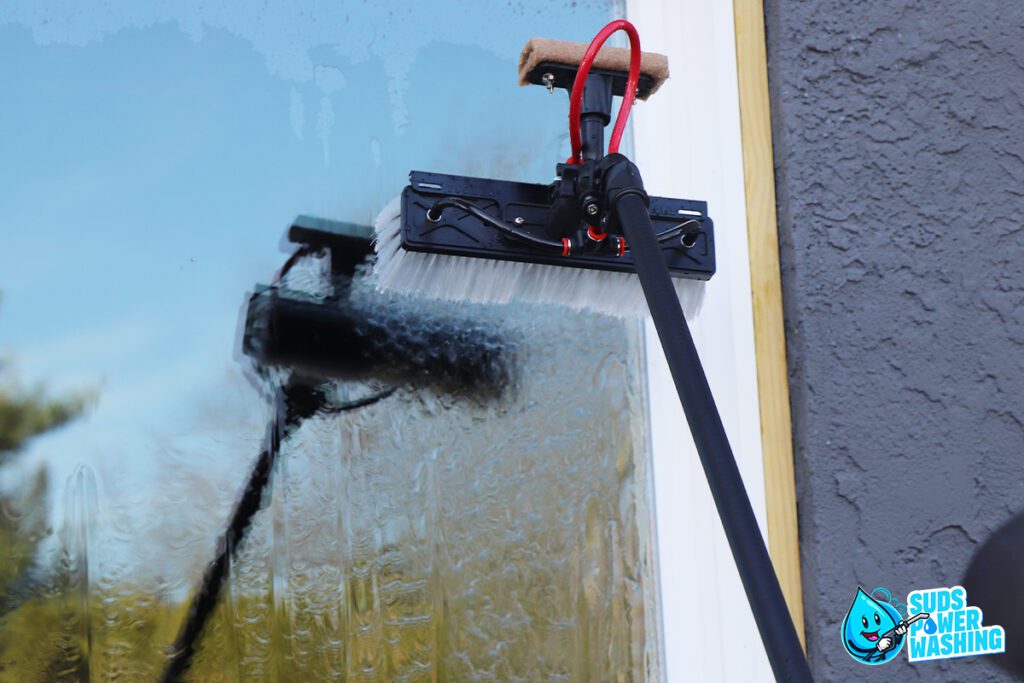 A close-up of a window being cleaned with a power washing tool. The commercial-grade tool has a brush attachment and water is being sprayed, creating a soapy surface on the window. The logo "Suds Power Washing" with a blue droplet mascot is visible in the bottom right corner.