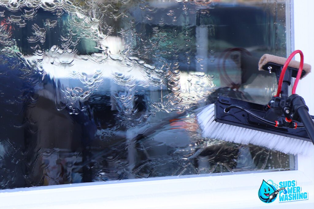 A window is being cleaned with a power washing tool, producing streams of water on the glass. The reflection of a person is visible in the background. In the bottom right corner, a blue character with the text "Suds Power Washing" is shown, highlighting their commercial cleaning expertise.