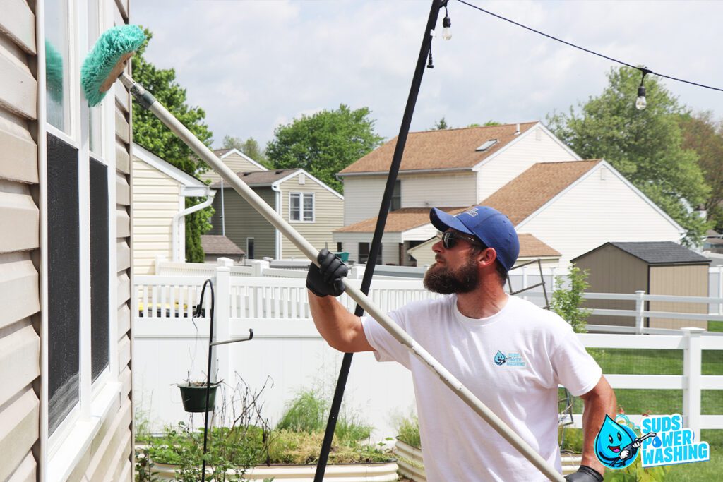 A person wearing a blue cap, white shirt, and gloves uses a long-handled brush to clean the upper window of a house. The background features houses, fences, trees, and outdoor lights. The "Suds Power Washing" logo is visible in the bottom right corner, highlighting their commercial services.