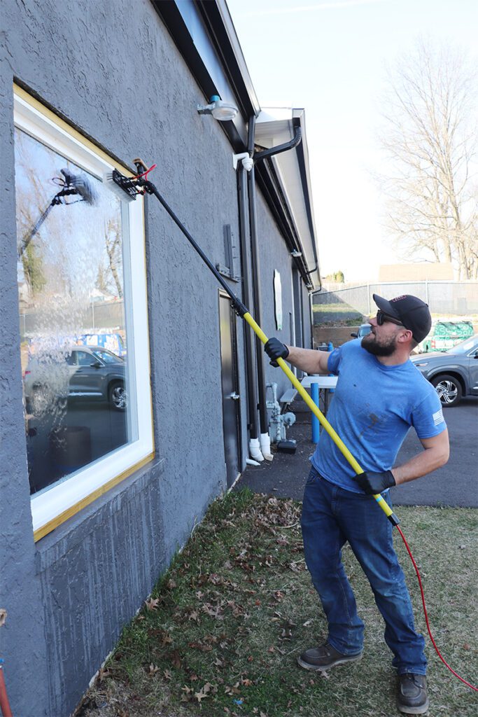 A man in a blue T-shirt, jeans, and a baseball cap is cleaning a large window on a gray commercial building. He uses a long pole with a brush attachment for the job. Wearing black gloves, he stands on grass next to the building, with trees and cars in the background.