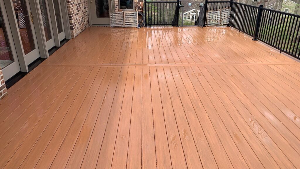 A large, freshly cleaned wooden deck is attached to a house with brick walls and multiple glass doors. The deck, reminiscent of a Davis Painting project, has black metal railings, beyond which a neighboring building and trees are visible. The deck's surface is wet, possibly from rain or cleaning.