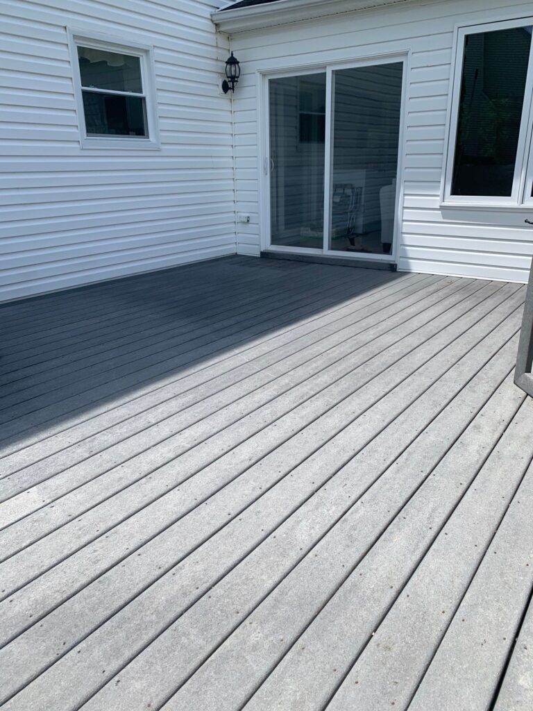 A wooden deck attached to a white house with horizontal siding showcases fresh residential painting. The deck extends from the exterior wall, featuring a sliding glass door and a window. An outdoor light fixture is mounted next to the sliding door, and the deck is partially sunlit.