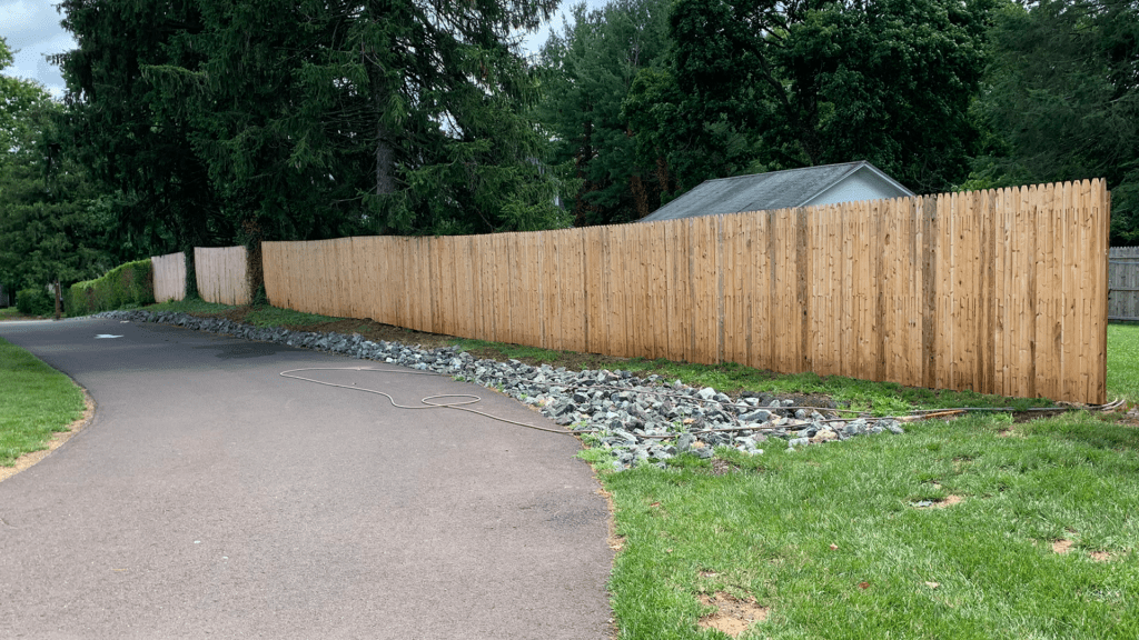 A long, tall wooden fence borders a paved path like a scene from a Davis painting. Rocks line the ground along the fence, and a coiled hose lies on the path. Trees and greenery fill the background, with a shed visible beyond the fence.