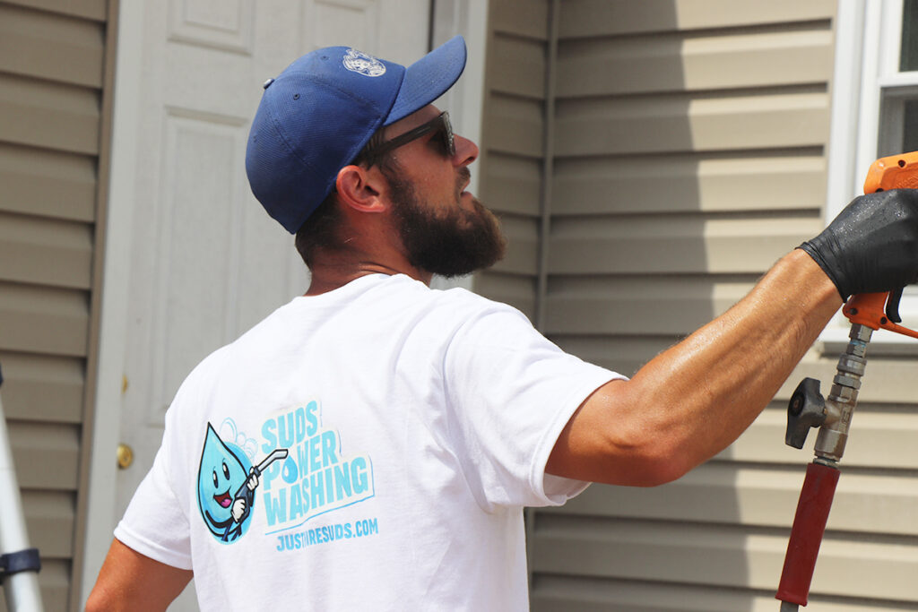 A man in a white "Suds Power Washing" T-shirt and blue cap uses a power washer to clean the siding of a house—showcasing one of the rewarding careers available in exterior cleaning. He wears sunglasses and a black glove on one hand.