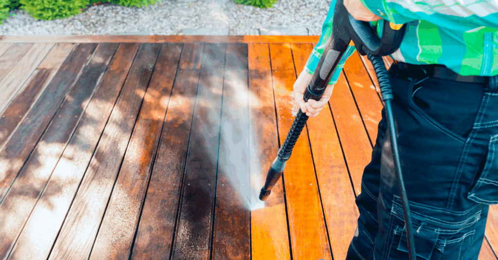 A person uses a pressure washer to clean a wooden deck, with a visible contrast between the cleaned and uncleaned sections of the wood.