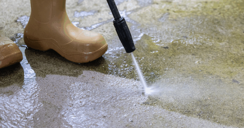A person wearing tan rubber boots uses a pressure washer to clean a dirty concrete surface, with water spraying powerfully from the nozzle.
