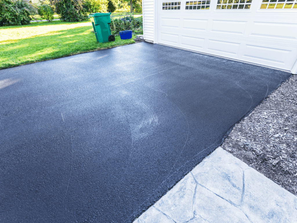 Freshly paved black asphalt driveway in front of a white garage, with green grass, a trash bin, and a blue recycling bin visible in the background. Sunlight highlights the smooth, clean surface.