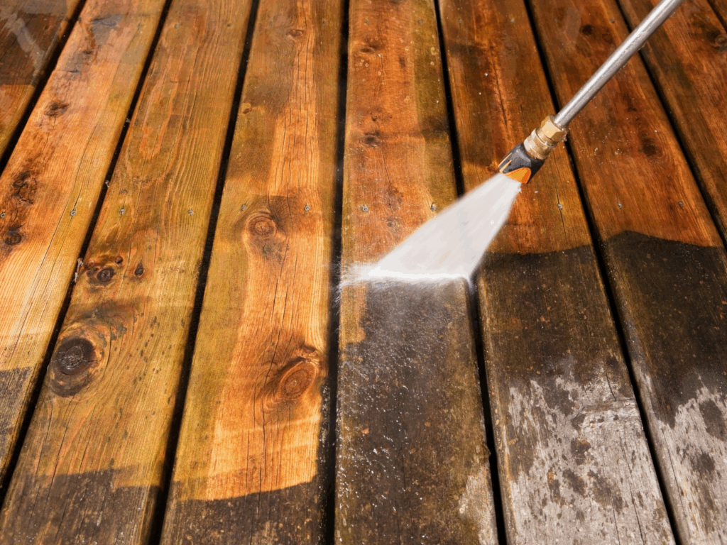 A pressure washer sprays water onto a wooden deck, cleaning away dirt and grime to reveal a lighter, cleaner surface compared to the surrounding unwashed areas.