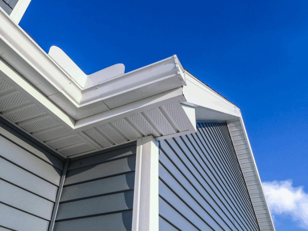 Close-up of a house corner with light blue vinyl siding, white soffit, fascia, and gutters under a clear blue sky. The clean lines highlight the modern exterior and roofing detail.