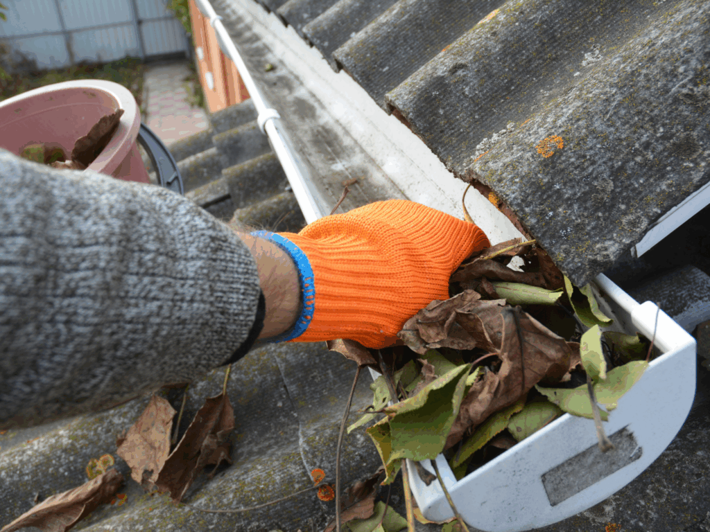 A person wearing an orange glove cleans out dry leaves and debris from a house gutter on a rooftop.