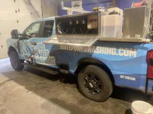 A blue truck labeled "Suds Power Washing" with equipment and tanks mounted in the bed, parked inside a garage. The website "IHatePowerWashing.com" is displayed on the side.
